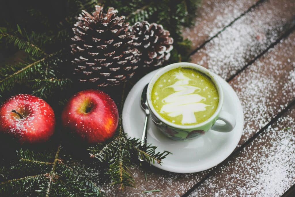 Cup of green matcha tee with motif of a Christmas tree on a wooden table with apples and pine cones.