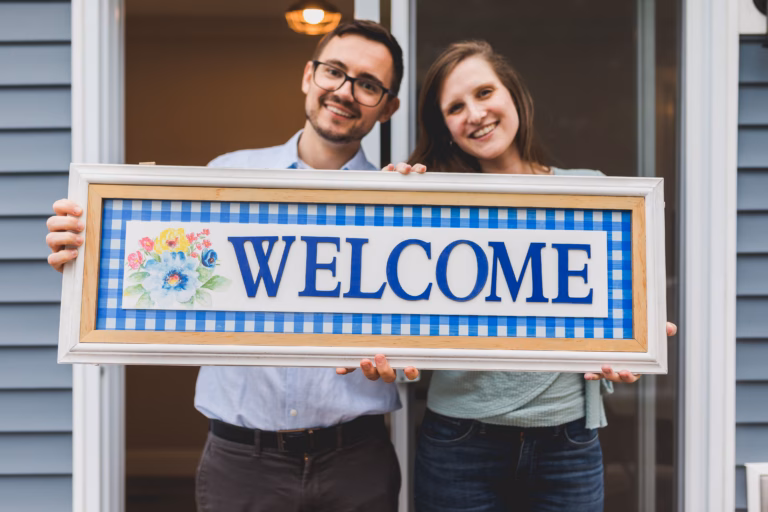 Airbnb-Ready: How to Organize Your Home for Short-Term Rentals in Vancouver and Victoria 3 A man and a woman, both smiling and holding a framed sign spelling "welcome". Illustration for Airbnb organization blog post.